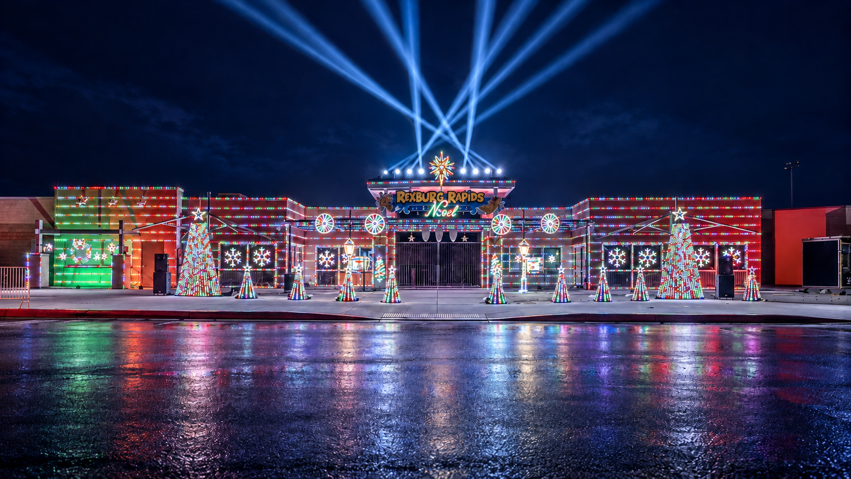 Noel at Rexburg Rapids illuminated in Christmas colors, with blue beams above the roof and reflections across the foreground.